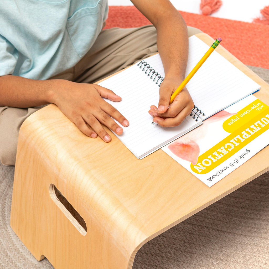 Child sitting on the floor using the Bentwood Floor Table, doing homework with a notebook and pencil on the spacious surface.