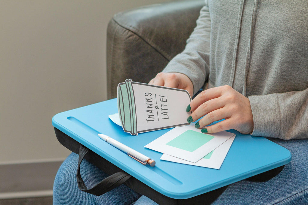 Person sitting in a chair using the Compact Lap Desk on their lap, writing on note cards with a pen on the smooth, portable surface.