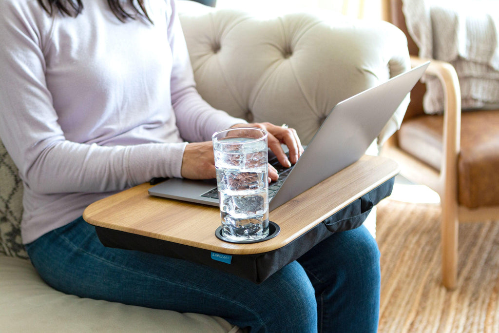 Person sitting on a couch using the Oakwood Cup Holder Lap Desk on their lap, working on a laptop with a beverage placed in the built-in cup holder.