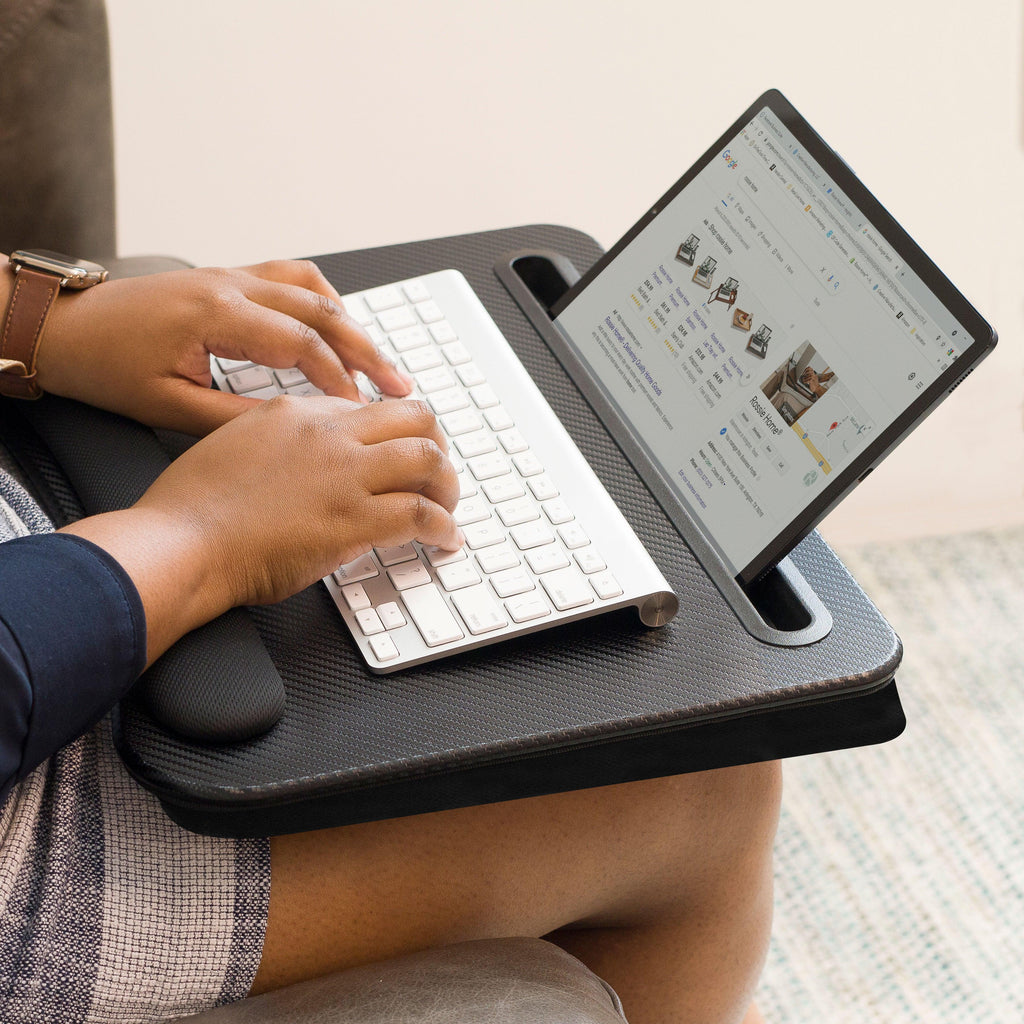 Lady sitting in a chair using the Black Carbon Memory Foam Lap Desk on their lap, typing on a Bluetooth keyboard with a tablet propped in the built-in media slot.
