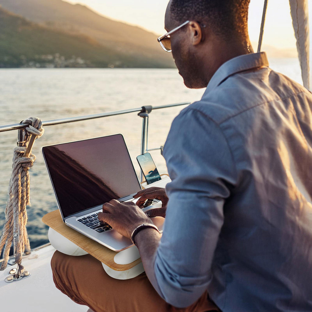 Person sitting on a boat with the White Oak Home Office Pro Lap Desk on their lap, working on a laptop with the built-in mouse pad, phone holder, and cushioned wrist rest for added comfort.